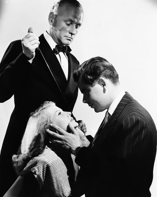 a black and white image of a man in a suit standing over a man and woman admiring each other on their knees